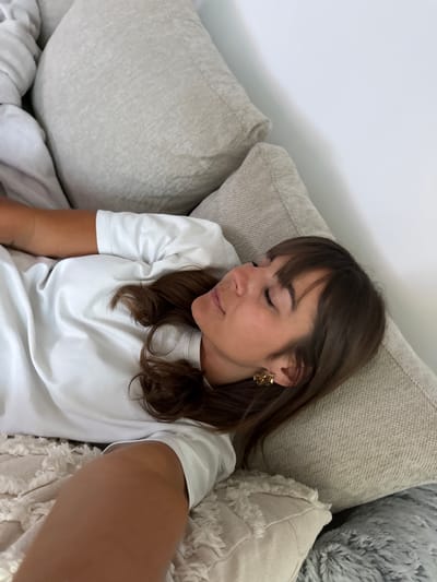 A serene, close-up shot captures a woman resting comfortably indoors, likely on a couch or bed. Her eyes are closed, suggesting she is sleeping or peacefully relaxing. She has medium brown hair with bangs, falling around her face and shoulders, and she wears a simple white t-shirt. A distinct gold earring with an organic or abstract shape is visible on her right ear. Her skin appears tanned, particularly on her visible arm.

She is surrounded by several soft, neutral-toned pillows, predominantly in shades of beige, cream, and light grey. Some pillows have visible texture, including one with fringed edges and another with a plush, fluffy texture. The background shows a plain white wall with a soft shadow cast on it, indicating indirect light. The overall atmosphere is tranquil and relaxed. No text is visible in the image. The setting, implicitly, is Cannes, France, though no specific visual cues of the location are present within the frame.