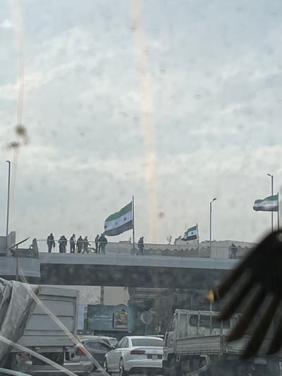The image displays an outdoor scene in Al-Muhajerin Municipality, Syria, captured from within a vehicle looking towards a concrete overpass or bridge under an overcast sky. Approximately 8-12 individuals are present on the overpass; most are standing in small groups near the center, while at least two figures are walking towards the right. Three flags, identifiable as the Syrian opposition/revolution flag (green, white, and black horizontal stripes with three red stars in the white band), are prominently displayed on poles along the overpass. Streetlights are visible on and adjacent to the overpass. Below the overpass, a road with vehicular traffic includes a white sedan and parts of two trucks. A large billboard featuring text and an image is visible beneath the overpass. The photograph contains numerous circular dust or dirt artifacts and a vertical light streak, suggesting it was taken through a soiled window or lens. A hand is partially visible in the lower right foreground, obscuring a portion of the scene.