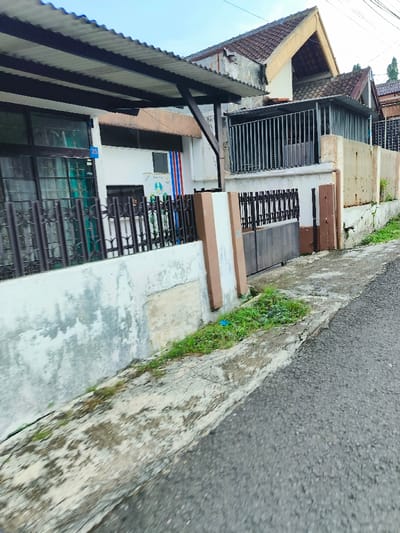 The image shows a street in Bandung, Indonesia, located at coordinates -6.895 latitude and 107.653 longitude. The street is paved with asphalt and runs along a white concrete wall with a brown concrete pillar. The wall has a metal fence on top of it and a small window. A brown wooden gate is on the right side of the wall. There is a small patch of grass in front of the gate. On the other side of the street is a building with a red tile roof. The building has a metal fence on the front and a wooden door. The image was taken from a perspective looking down the street. There are no people in the image.