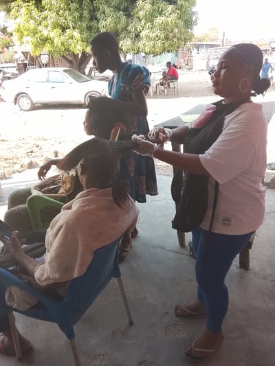 The image depicts an outdoor or semi-outdoor scene during bright daylight, likely a hair salon or styling area. In the foreground, three individuals are engaged in a hair styling activity.
On the far left, seated in a blue plastic chair with their back to the camera, is a person whose hair is being treated. They are draped with a light-colored (possibly beige or light brown) towel or shawl over their shoulders. Their long, dark hair is pulled back and sectioned, heavily coated with a thick, greenish-brown product, suggesting a chemical treatment like a relaxer, dye, or deep conditioner. This person holds a mobile phone in their left hand, appearing relaxed while waiting. Behind them, several stacked tires are visible, possibly serving as additional seating or part of the outdoor setup.
Standing to the right of the seated individual and facing left is a woman actively working on the hair. She wears a white short-sleeved t-shirt with small text visible on the front (partially readable as "UNLIMITED"), blue leggings, and light-colored flip-flops. She has a dark apron or bib covering her front and is wearing clear plastic gloves on both hands as she applies the product to the client's hair with focused attention. Her hair is pulled back, and she wears large hoop earrings.
Between the seated client and the standing stylist, a young person, possibly a child or teenager, stands observing or assisting. They are wearing a blue patterned dress and have short, dark hair, looking down at the hair being worked on.
In the mid-ground, a white car is parked on the left side, partially obscured by a large, leafy green tree. Further in the background on the right, another person in a red top is visible seated at a table, along with other blurry figures, suggesting a bustling outdoor environment. The ground is a light-colored concrete or paved surface, showing some wear and cracks in places. The bright sunlight indicates a clear day, casting soft shadows.