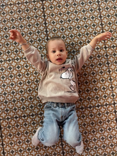 A baby wearing a beige sweater with Snoopy on it and jeans is lying on a patterned tile floor. The baby is lying on their back with their arms outstretched to their sides. The background is a tile floor with a pattern of black and white crosses on a brown background.