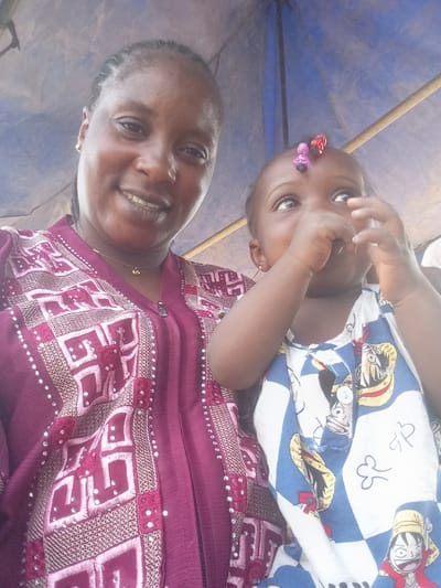 A medium, eye-level, full indoor shot depicts an adult woman and a baby together. The woman is light-skinned with dark braided hair, wearing a maroon patterned outfit with a zipper and a delicate gold necklace. She smiles into the camera. The baby, wearing a blue and white patterned top featuring cartoon characters, looks upward with their fingers near their mouth, and has colorful hair clips. The setting appears to be under a blue canopy or umbrella. The image was taken in Doma, Nigeria.