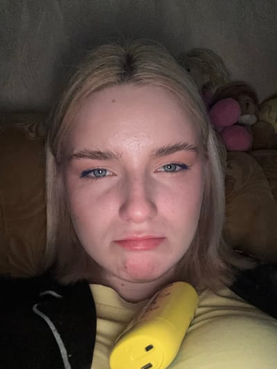 The image is a close-up, vertically oriented, selfie-style photograph of a young person with blonde hair. The person is lying down, and their face is tilted towards the camera. They have light blue eyes and are wearing dark blue eyeliner. Their lips are slightly parted, and their skin appears to be pale with some subtle redness on the cheeks.
In the background, there are what appear to be stuffed animals, including one with pink pom-pom-like features and another that is brown and fuzzy. To the right of the person's face, a bright yellow cylindrical object is visible, which might be a bottle or a portable charger, with what looks like USB ports. The person is wearing a light yellow garment on their upper body. The lighting is dim, suggesting it might be nighttime or a poorly lit room. The overall mood appears relaxed or casual. There is no visible text in the image.