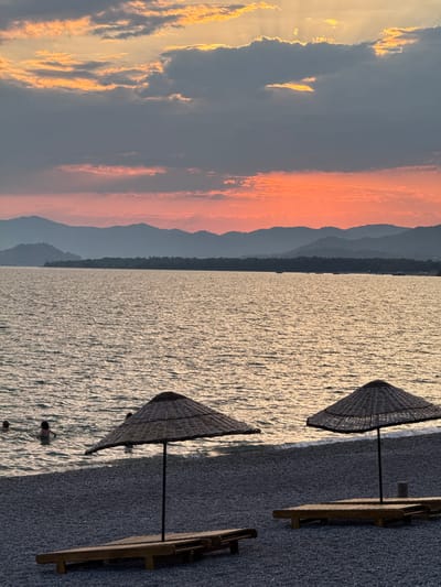 This image depicts a serene sunset scene at a pebble beach in Fethiye, Turkey. The sky is ablaze with the vibrant hues of orange and pink, with sunbeams piercing through scattered clouds, creating a dramatic backdrop. In the foreground, two thatched beach umbrellas stand over empty wooden lounge chairs, suggesting a peaceful end to the day. The calm sea gently laps at the shore, and in the distance, a line of silhouetted mountains adds depth to the landscape. A few individuals are visible in the water, enjoying the last moments of daylight, their presence adding a touch of life to the tranquil setting. The overall atmosphere is one of quiet beauty and relaxation.