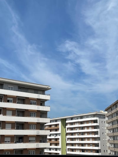 The image shows a view of apartment buildings against a blue sky with wispy clouds. The buildings are modern, with multiple stories and balconies. The color palette is dominated by neutral tones of white, beige, and brown, with a vertical strip of green visible on one of the buildings. The scene appears to be a residential area within a city, potentially Podgorica, Montenegro, characterized by urban architecture and a clear sky. There are no people or specific actions depicted, focusing solely on the architectural structures and the atmospheric conditions.