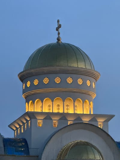 The image shows the top of the Church of St. John Vladimir in Bar, Montenegro, at dusk. The church features a prominent green dome topped with a cross, surrounded by decorative windows with intricate patterns illuminated from within. Below the dome, there is a band of arched windows also glowing with warm light. The building itself is white with sharp architectural details, including angled roof sections with downward-facing lights. A portion of another dome, also with a glass-like surface, is visible in the foreground. The sky is a deep, clear blue, indicative of the twilight hours. There are no people visible in the image.