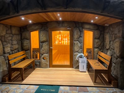 This is an eye-level, exterior medium shot of a modern sauna facility located in Ban Bang Rak, Thailand. The facility is built into a rock formation, with a wooden ceiling and deck. The entrance to the sauna is a glass door, revealing a well-lit interior with wooden benches. On either side of the entrance are two windows, each looking into separate chambers with what appear to be heating elements. Two wooden benches are positioned outside the sauna, one on each side. A white humidifier with a silver flexible hose is situated in front of the sauna door. A green "WELCOME" mat lies on a stone pathway in front of the wooden deck. The overall impression is one of a clean and inviting spa environment, designed to blend with a natural setting.