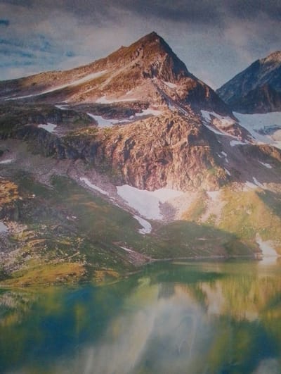 The image depicts a vast, mountainous landscape with a serene body of water in the foreground. The mountains are rugged and rocky, with patches of snow clinging to their slopes. Sunlight illuminates some of the peaks, casting shadows and highlighting the textures of the rock. The sky above is a mix of blue and gray clouds, suggesting a dynamic weather pattern.
The water in the foreground is calm and reflective, mirroring the surrounding mountains and sky. The colors in the water range from deep blues and greens to lighter shades, creating a tranquil and almost ethereal effect. The overall scene evokes a sense of natural beauty and solitude.
There are no people, animals, or man-made structures visible in the image. The environment appears to be a remote and undeveloped natural area.
This image does not depict the city of Kasumkent, Russia, nor any part of it. The landscape shown is a natural mountain environment and does not contain any urban elements or recognizable landmarks of Kasumkent.