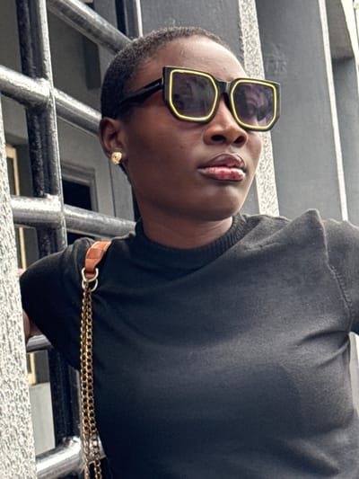 A dark-skinned person wearing dark sunglasses and a black sweater is the primary focus of the image. They appear to be leaning over a metal railing, possibly of a staircase or balcony, with a building or structure visible in the background. The subject is wearing a gold earring and a brown leather belt with a gold chain detail. The perspective is from below, looking up at the individual. The location is likely Ojomu, Nigeria.