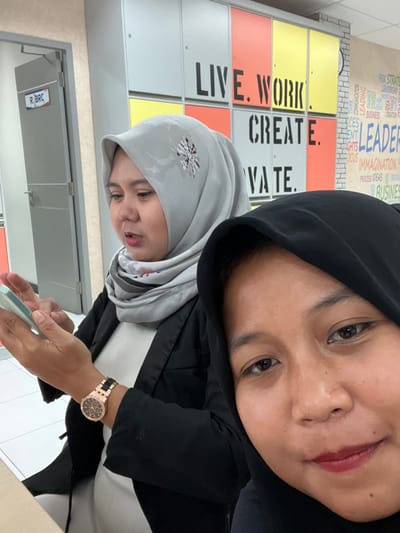 The image shows two women, both wearing hijabs, in an indoor setting. The image is rotated 90 degrees clockwise, making the women appear tilted to their left.
In the foreground, a woman with warm-toned skin is captured in a close-up, facing towards the right side of the frame (when oriented correctly). She is wearing a black hijab that frames her face, revealing her forehead and part of her cheeks. Her eyes are open and looking slightly upwards, and she has soft, natural makeup with reddish lipstick. She is wearing a dark top, possibly a jacket, over a white inner layer. A rose gold-toned watch with a black band and a crystal-studded bezel is visible on her left wrist. Her right hand is holding a light-colored smartphone or similar device.
Behind the foreground woman, slightly to her left, another woman is partially visible. She has light-toned skin and is wearing a light grey hijab that features a circular, stylized pattern resembling a snowflake or starburst in black, white, and red on the side near her temple. Her mouth is slightly open as if she is speaking or reacting to something. She is looking in the direction of the foreground woman.
The background consists of a wall with a grid of colorful panels, primarily light yellow, orange, and light grey. Large, stenciled black text is written across these panels, forming motivational words. Above these large words, smaller, lighter text is also visible on a white brick-patterned section of the wall. Below the women, a light-colored floor is visible.
To the left of the women, a closed door is visible with a sign that reads "R. BRC".
Visible text on the wall includes:
- "LIVE. WORK"
- "CREATE."
- "VA TE." (The first letter might be obscured, possibly "INNOVATE" or "CULTIVATE")
- "LEADERSHIP"
- "BUSINESS"
- "IMAGINATION"
- "PROCESS IDEAS"
- "BUSINES" (partially visible)
- Other smaller words like "STRATEGY," "COMMUNICATION," "FOCUS," "GOALS," "INSIGHTS," "DREAM" can be vaguely seen.
The overall atmosphere appears casual, possibly in an office or educational environment given the motivational wall art and general setting. Both women seem engaged, with the foreground woman focused on her device and the background woman possibly conversing.