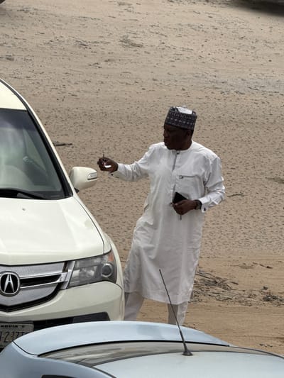 A medium-shot image shows a man in a white tunic and patterned cap standing by a white SUV. He holds a key near the vehicle's side mirror. The car's license plate reads "Abuja." The location appears to be a street or parking area in Lugbe, Nigeria.