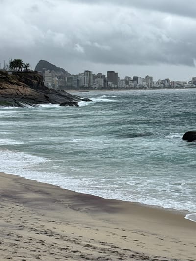 The image presents a wide view of a coastal city under an overcast sky, showcasing the interface between urban development, natural rock formations, the ocean, and a sandy beach.
On the left side, a prominent, dark rocky outcrop extends into the water, with several palm trees visible near its base. Behind this, a dense skyline of numerous high-rise buildings stretches horizontally along the coastline, indicating a significant metropolitan area. The buildings, mostly light in color, rise several stories high, creating a busy urban backdrop.
The middle section of the image is dominated by the ocean, characterized by light grey-blue water. Numerous waves are actively breaking, creating extensive white foam as they advance towards the shore, suggesting a moderately active sea.
To the right, a broad sandy beach occupies the foreground. The sand exhibits a light brown hue with darker, damp patterns near the waterline. Multiple footprints are visible across the sand, indicating recent human activity, although no people are present in the frame. The boundary where the foamy waves meet the sand is distinct.
The sky above is uniformly overcast, filled with light grey clouds, suggesting a cloudy or potentially rainy day. The lighting is soft and diffuse, lacking strong shadows, which points to daytime conditions, likely late morning or afternoon. The combination of a mountainous, rocky coastline, extensive beach, and a high-rise urban environment strongly aligns with the geographical characteristics of Rio de Janeiro, Brazil. There is no visible text in the image.