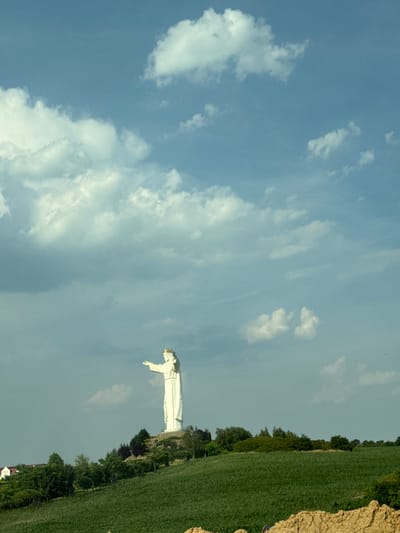 The image shows a large, white statue of Jesus Christ, with its arms outstretched. The statue is situated on a hilltop, overlooking a verdant landscape of green fields and trees. The sky is a pale blue, with scattered white clouds. The scene appears to be set in a rural area, likely in Poland given the location context. The time of day seems to be late afternoon or early evening, with the sun casting a soft light on the scene. There are no people visible in the immediate vicinity of the statue, and no text is present in the image. The overall mood of the image is one of peace and serenity.