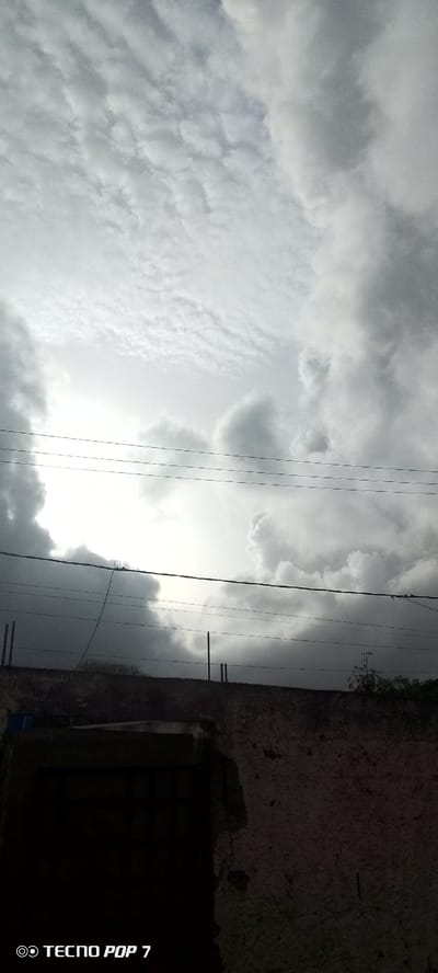 The image shows a cloudy sky over a wall with electrical wires. The clouds are a mix of grey and white, with some areas of bright light suggesting the sun is behind them. The wall appears to be made of concrete or brick and has some texture. Electrical wires are strung across the frame, running horizontally. There are also some vertical poles visible along the wall. The bottom left corner of the image contains a watermark that reads "TECNO POP 7". The scene appears to be outdoors, possibly in a residential or urban area. The overall impression is one of an overcast day.