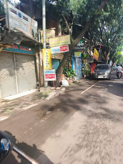The image shows a street in Bandung, Indonesia (latitude: -6.901, longitude: 107.647) with a closed garage door and several shops on the left side of the frame. There are multiple signs in Indonesian and English for tailors and a bakery. A large tree trunk is visible in the center of the frame, with a white bag lying beneath it. There is a gray car parked on the right side of the street, facing the camera. A motorcycle is visible at the bottom of the frame, seemingly taken from the perspective of the rider. The street is asphalt and the day is sunny. The image is taken from a moving vehicle.