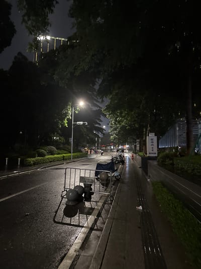 The image depicts a nighttime street scene in Haizhu District, China, likely after rainfall, as the asphalt road and sidewalk appear wet and reflective. The scene is illuminated by a streetlamp and the lights of a building in the distance. On the right side of the road, a modern building with illuminated vertical elements is visible, adjacent to a landscaped area. A sign with Chinese characters is placed in front of this building. A sidewalk runs alongside the road, and further down, several parked motorcycles are visible. In the foreground, a metal barricade is set up on the side of the road, with two dark, spherical objects resting on the ground behind it. Trees with dense foliage overhang the street and sidewalk, creating a dark, shadowy atmosphere. The road itself is mostly empty, with only a few distant vehicles suggesting the presence of traffic.