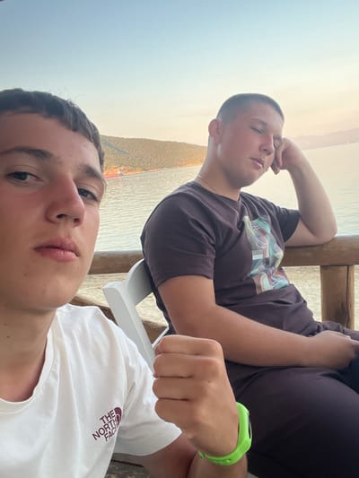 The image features two young men in a selfie taken outdoors. The man in the foreground, on the left, is close to the camera, showing his face from the nose up. He has dark hair and is wearing a white t-shirt with "THE NORTH FACE" logo visible on the right side of the chest. The second man, to the right and slightly further back, is lying down, resting his head on his arm. He has short hair and is wearing a dark t-shirt with a graphic print and a bright green watch on his left wrist. Both individuals appear relaxed.
The background suggests a waterside setting, likely a beach or coastline, with calm water and a hazy, possibly warm, sky indicating it is either early morning or late afternoon. The lighting is soft and diffused. They are seated on what appears to be a wooden structure, possibly part of a bench or a deck overlooking the water. There are wooden beams visible in the background, and a white chair is also present. The scene evokes a sense of leisure and vacation. The location context is Nea Peramos, Greece.