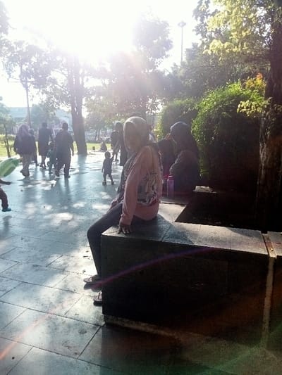 The image shows a young woman sitting on a concrete ledge. She is wearing a hijab and a pink jacket. She is looking towards the right side of the image. To her right is another person sitting on the same ledge. The person is wearing a dark jacket and their back is towards the camera. The image appears to be taken in a public park in Pati, Indonesia (latitude: -6.754, longitude: 111.04).  There are people walking around in the background and a young child is walking towards the left of the image. The image is slightly backlit with the sun shining through trees in the background.  The ground is made of concrete tiles.  There are some green plants and trees around the scene. The image is taken from a slightly elevated perspective, looking down on the scene. The lighting is bright, and the colors are muted. The focus is on the young woman in the foreground, and the rest of the scene is blurred.  The composition of the image is balanced, and the overall effect is serene.