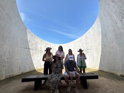 This image captures six individuals within the architectural structure known as the Cuernavaca Cathedral of Light (Sunstone), located near San Isidro Llano Grande, Morelos, Mexico. The scene is enclosed by a tall, curved, light grey concrete wall, open to the clear blue sky above. The ground within the structure consists of sand and sparse green vegetation bordering the wall. In the foreground, a dark, long bench is positioned centrally. A male subject is seated on the bench to the right, wearing a striped polo shirt, white shorts, a white baseball cap, and flip-flops, with his head down. To his left, a female subject is seated on the bench, wearing a long patterned dress and a straw hat. Standing behind the bench are four additional female subjects: one on the far left wearing a light tank top, dark pants, and a wide-brimmed hat; two in the center with light tops, one with sunglasses on her head, and the other with sunglasses; and one on the far right wearing a white sheer cover-up over green shorts and a wide-brimmed hat. Sunlight casts distinct shadows on the right side of the interior wall.