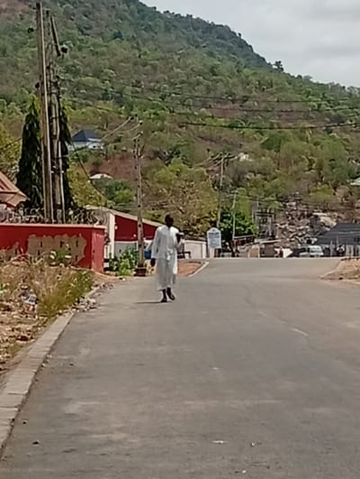 The image displays a daytime street scene in Adankolo, Nigeria. A paved road occupies the foreground and midground, curving slightly to the right in the distance. A lone individual, dressed in a long white garment and dark shoes, walks away from the viewer on the left side of the road, approximately centered in the frame. To the immediate left, a concrete curb borders a narrow dirt strip containing dry vegetation and scattered refuse. Beyond this, a red-painted wall with an patterned top edge runs parallel to the road, partially obscuring unseen structures. Multiple wooden utility poles, strung with overhead power lines, are present on both sides of the road, prominently on the left. The background is dominated by a large, green, tree-covered hill or mountain, with several residential buildings visible on its lower slopes. In the far distance along the road, vague outlines of additional buildings and vehicles are discernible. A white signpost is visible on the right side of the road in the mid-distance. The sky is partially cloudy.