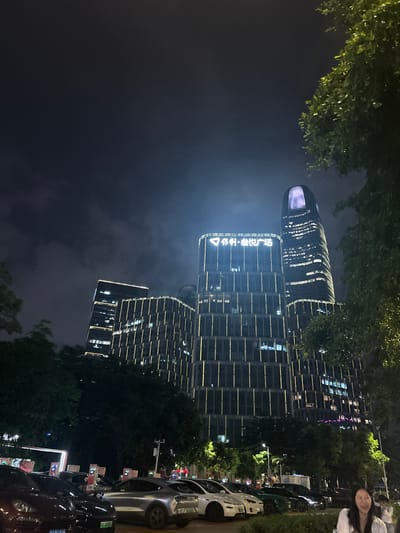 This media file captures a nighttime scene in Haizhu District, China, featuring a modern architectural landmark. The primary subject is a tall, multi-story building with a distinctive curved top that glows with internal lighting. The building's facade is a grid of windows, some illuminated, creating a striking visual against the dark sky.
In the foreground, lush green trees with dense foliage frame the top and right side of the image, their leaves illuminated by the ambient light. The lower part of the scene shows several parked cars, including a sleek silver car and a darker vehicle. The overall setting is urban, with streetlights and the building's illumination suggesting a well-developed commercial or residential area.
A woman is partially visible on the far right, smiling at the camera, adding a human element to the scene. Her presence suggests a casual, perhaps social, gathering or outing. The time of day is clearly night, indicated by the darkness of the sky and the prominent artificial lighting. The weather appears calm, with no visible signs of rain or strong winds.
Visible text includes Chinese characters on the building that read "科创·金融城" (Ke Chuang · Jin Rong Cheng), which translates to "Science and Technology Innovation · Financial City." License plates on the cars are also visible, though the characters are too small to be legible. The lighting, the modern architecture, and the presence of a financial district sign point to a developed, contemporary urban environment in China. The image evokes a sense of urban vibrancy and modern progress.