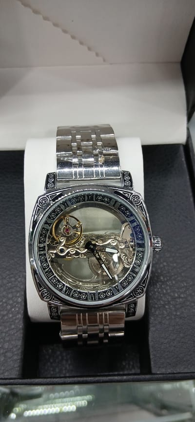 A mechanical wristwatch with a silver-toned metal band is displayed on a white stand. The watch face is transparent, revealing the inner workings of the timepiece, including gears and springs. Roman numerals mark the hours on the bezel, which is adorned with an intricate, dark pattern. The watch is positioned in the center of the frame, with a blurred black surface visible in the foreground and background. The environment appears to be a display case or retail setting, possibly within Akmachit, Uzbekistan, though this specific location cannot be confirmed solely from the visual information provided. The lighting is bright and even, highlighting the metallic sheen of the watch.