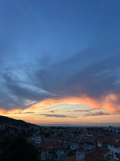 The image captures an expansive, high-angle view of the city of Asenovgrad, Bulgaria, during a sunset. The sky is a dramatic canvas of deep blues and grays, transitioning into vibrant hues of orange and pink along the horizon, where the sun is setting. Wispy clouds are scattered across the sky, some catching the last rays of sunlight and glowing with warm colors. Below the sky, the cityscape unfolds. The foreground is dominated by the rooftops of numerous buildings, characterized by their terracotta tiles and a mix of architectural styles. A dense collection of trees, dark green and silhouetted against the fading light, occupies the left side of the frame, obscuring parts of the hillside and buildings. The city extends towards the background, with more buildings and structures visible, interspersed with glimpses of distant, hazy mountains. The overall atmosphere is serene and picturesque, indicative of the end of the day.