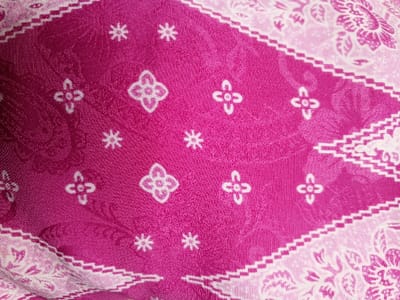 A close-up captures a pink and white textile with a patterned design. The main section is a vibrant pink, adorned with small, repeating white floral and star-like shapes. Framing this is a white border with a delicate floral pattern and a zigzag edge. The fabric appears to be folded, creating a textured and slightly obscured view of the overall design.