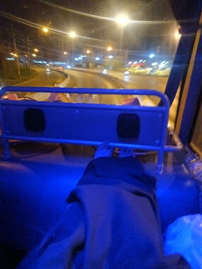 The image appears to be taken from inside a vehicle, possibly a tuk-tuk or similar transport, at night in Hua Hin City Municipality, Thailand. The street is visible through the front window, with street lights and blurred headlights of other vehicles. A blue metal frame with a cushioned back is in the foreground. In the bottom of the frame, a person is lying down covered in a grey blanket. A plastic bag can be seen on the right side of the frame. The overall lighting is blue-toned.