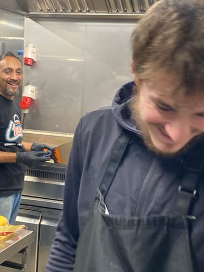 Two men are visible in a commercial kitchen. The man closer to the camera, partially cropped, has light hair and a beard, wearing a black hooded sweatshirt and apron. The other man is wearing a black t-shirt and black gloves. He smiles and holds a small orange and black tool. Behind them, are stainless steel walls and equipment, with visible electrical outlets and ventilation. The scene is brightly lit, suggesting a functional workspace.
