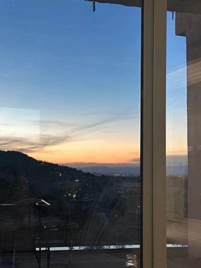 The image captures a sunset view from a window overlooking a hilly landscape with scattered lights, possibly indicating a town or city. The sky displays a gradient of colors, transitioning from deep blue at the top to orange and yellow near the horizon. A faint wispy cloud formation is visible in the upper sky.
In the foreground, the window frame is visible on the right side of the image, and a reflection of what appears to be an interior space, including a light fixture, can be seen on the glass. On the left side of the image, another reflection is present, showing a dark object that might be a table or railing, and a portion of a hand holding a phone.
The landscape below is characterized by a dark, silhouetted hillside dotted with numerous small lights. This suggests a residential or urban area situated on the slopes. In the distance, the horizon is marked by faint lights, hinting at a larger settlement. A tall, slender structure with lights on it, resembling a communication tower or a prominent building, is visible on the hillside. The overall scene conveys a tranquil evening atmosphere.
Considering the location of Asenovgrad, Bulgaria, this view likely corresponds to a vantage point within or on the outskirts of the city, offering a panorama of the surrounding hills and the city lights spreading across the valley. The presence of lights and structures on the hillside strongly suggests the urban environment of Asenovgrad.