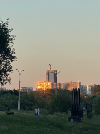 The image depicts a panoramic view of the city of Minsk, Belarus, at sunset. In the foreground, a grassy field is dotted with patches of wildflowers. To the left, a park bench is visible, while a woman in a white top and blue jeans stands in the center of the field. To the right, a man in a green jacket and dark pants walks away from the camera. In the mid-ground, a thick line of trees separates the park area from the urban landscape. Beyond the trees, several buildings are visible. A tall, unfinished building with a crane on top stands prominently, with a large, multi-story apartment building behind it, reflecting the warm hues of the setting sun. A street lamp with two lights is positioned to the left of the tall building. A dark, abstract sculpture composed of vertical metal elements is in the right mid-ground. The sky is a soft gradient of pale blue to warm orange, indicating the time of day.