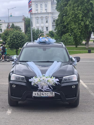 A black BMW X3 SUV, decorated for a wedding, is centrally parked on asphalt pavement. The vehicle features an arrangement of white and light blue artificial flowers with light blue sheer fabric ribbons draped over the front hood. A smaller floral arrangement, also in white and light blue, sits on the roof, topped with two interconnected golden rings. Small clusters of similar flowers are affixed to the side mirrors. The front license plate displays "X624TC 18 RUS".
In the background, under an overcast sky, the urban environment of Sarapul, Russia, is visible. To the far left, two partially obscured individuals on bicycles are present, with one wearing a helmet and backpack. Behind them, green foliage partially conceals a building with a grey roof and a prominent green onion dome, suggesting a religious structure. A smaller flag with Russian national colors is faintly visible near this building. Towards the center-background, a tall, slender pole displays a spiraling ribbon in the white, blue, and red colors of the Russian flag. Adjacent to the pole stands a stately, light-colored, multi-story building with numerous windows and classical architectural details. To the right, large mature green trees are present, along with a dark cylindrical waste bin. The ground is asphalt with faded white parking lines visible on the right.