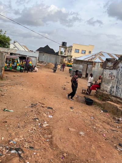 The image depicts a street scene in Barakin Akawo, Nigeria. A dirt road is covered with debris, including plastic bags, wrappers, and scattered rocks. On the left side of the road, a yellow and green auto-rickshaw is parked. Several people are gathered around it, some standing and some seated. Further down the road, a man pushes a wheelbarrow.

In the background, corrugated metal structures and buildings with zinc roofs are visible. One building has a light yellow facade with blue-framed windows. The sky is overcast with clouds.

Several individuals are present in the foreground and midground. A young man in a black basketball jersey with the number "2" walks towards the left of the frame. To his right, a boy in a white t-shirt and patterned pants is speaking to a woman in a red skirt and headscarf, who is seated on a raised concrete structure. A black bucket is placed on the ground near them. Another group of people are congregated near the auto-rickshaw.