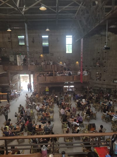 The image shows the interior of a large, industrial-style space filled with people seated at numerous small tables. The setting is a multi-level structure with exposed beams, brick or concrete walls, and several windows admitting natural light. On the upper levels, people are seated along balconies or walkways overlooking the main floor. Red banners with Chinese characters are visible hanging within the space. Individuals are engaged in various activities, primarily eating, drinking, and socializing. The seating arrangement is dense, indicating a crowded or busy environment. The image suggests the scene is in the city of Liangjiang, China.
