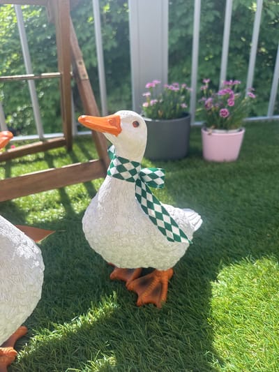This image depicts a decorative goose statue placed on a patch of artificial grass. The goose is white with an orange beak and eyes. It wears a green and white checkered ribbon tied around its neck. The statue's feet are visible, also orange. The background suggests an outdoor setting, possibly a balcony or patio, with a railing and greenery visible. Two potted plants with small purple flowers are positioned behind the goose, one in a pink pot and another in a dark gray pot. The lighting indicates it is daytime, with sunlight casting shadows on the grass. Another goose statue, partially visible at the bottom right of the frame, suggests this is part of a garden decoration. No text is visible in the image. The overall mood is cheerful and decorative.