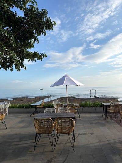 The image depicts an outdoor seating area overlooking a body of water. A large tree with dense green foliage is visible in the upper left corner, casting shadows on the tiled ground. Several tables and chairs, made of woven material with metal legs, are arranged on the patio. Some tables have chairs around them, while others are empty. In the background, a white umbrella stands near the water's edge. Beyond the patio, a shoreline with dark pebbles and a strip of green plants separates the seating area from the water. The water is calm, with gentle ripples extending towards the horizon. Two small figures are visible in the shallow water, and a boat is anchored further out. A wooden swing set structure is present near the water. The sky is blue with scattered white clouds. The scene appears to be a tranquil outdoor dining or lounging area.