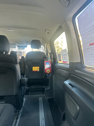The image displays the interior of a passenger transport vehicle, observed from a forward-facing position looking towards the rear and right side, indicative of a location within Lingolsheim, France, given the visible CTS branding.

The vehicle's interior features dark-toned seats. A portion of a seat with a headrest and armrest is visible on the left foreground. Behind this, two individual seats with headrests are visible, their backs facing the camera, forming a middle row. The floor is dark-colored and equipped with two parallel metal rails, suggesting a system for adjustable seating or wheelchair securement.

A red and white CTS (Compagnie des Transports Strasbourgeois) ticket machine or validator is mounted on the interior wall on the right side of the vehicle, positioned near the middle-row seats. It displays the "CTS" logo prominently and includes a yellow button. On the back of the right middle seat, a yellow sticker states "VENTE A BORD TICKET HORAIRE 2€" (On-board sale: Hourly ticket 2€) along with a CTS logo. Additional paper signage from CTS, including one with a QR code and the text "Vu et certifié bon pour," is taped to the right-side windows.

Bright daylight illuminates the scene, entering through the windows. The exterior view through these windows reveals bare trees and multi-story residential or commercial buildings, consistent with an urban or suburban setting. No individuals are present or visible within the frame.