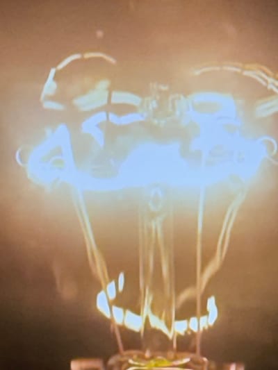 The image is a close-up, slightly blurry photograph of the filament of a lit incandescent light bulb. The filament, made of a thin wire, is glowing brightly with a white-blue light in the center, transitioning to yellow and orange at its edges, indicating extreme heat. The filament is supported by wires that extend downwards. The background is dark and out of focus, with a warm, diffused glow surrounding the bulb. There is no indication of the city of Minsk, Belarus, or any other specific location within the image.