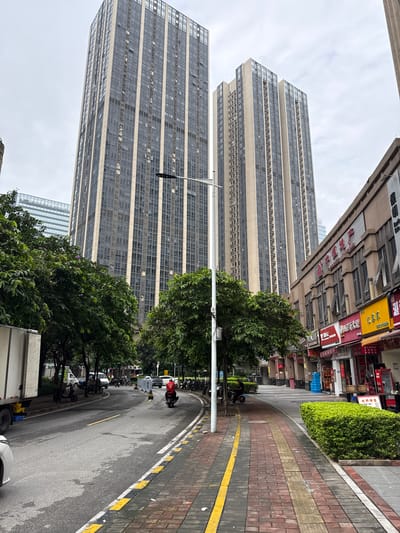The image depicts a street scene in Haizhu District, China, dominated by towering residential buildings with glass facades. A paved road curves through the scene, with a white truck on the left and several motorcycles parked along the roadside. A person on a motorcycle wearing a red jacket is visible on the road. A sidewalk, paved with red and yellow bricks, runs alongside the road, lined with lush green bushes. A row of commercial establishments with awnings and signage is situated on the right side of the sidewalk. Several trees with full green foliage are present, casting shadows on the street. The sky is overcast, suggesting a cloudy day.