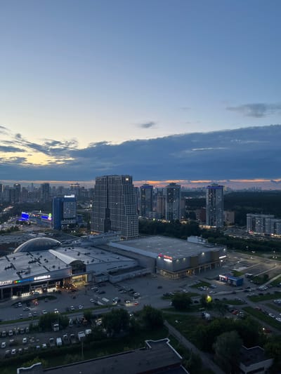 This is an aerial, high-angle, dusk shot of a modern urban landscape, likely in Yekaterinburg, Russia, given the Russian text visible on some buildings. The image captures a complex of large commercial buildings, including a shopping mall with a distinctive domed section and a large, brightly lit storefront. In front of these buildings, a busy parking lot is filled with numerous cars.
Beyond the commercial area, a dense forest stretches into the distance, bordering a cityscape dominated by numerous high-rise residential and office buildings. Several of these skyscrapers are illuminated, with some featuring blue accent lighting on their upper floors. The sky is a gradient of twilight colors, transitioning from a pale blue near the horizon to a deeper, cloud-streaked blue overhead.
The overall scene depicts a developing area of a Russian city, with a clear demarcation between commercial/residential zones and natural green spaces. There are no discernible individuals or specific actions taking place that are significant enough to detail. The focus is on the urban development and the natural environment interacting.