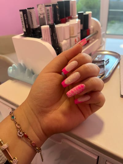 The image shows a close-up of a person's left hand, displaying brightly colored acrylic nails with a French tip design. The nails are long and have a squared-off shape. The nail polish colors include hot pink and white with glitter. The hand is resting on a white surface, possibly a vanity or desk, with a blurred background that appears to be a nail salon. In the background, various nail polish bottles and salon equipment are visible, arranged in a white organizer. A delicate bracelet with colorful charms is worn on the wrist. The overall impression is of a professional nail service being showcased. There is no indication of the city of Novi Pazar, Bulgaria in the image.