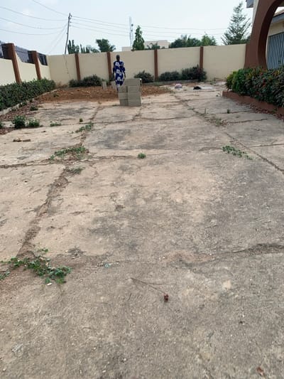 A wide view of a cracked concrete ground interspersed with weeds and some plant life growing near a wall. An individual stands next to a stack of grey bricks. The scene gives the impression of a neglected or undeveloped outdoor space. The sky is overcast. Buildings are partially visible along the left edge of the scene.