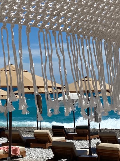 The image depicts a beach scene in Prijevor, Montenegro, viewed through a decorative white macrame fringe. In the foreground, several empty lounge chairs made of wood and tan fabric are arranged on a pebble beach. Some chairs are partially covered by beach umbrellas, which are tan and white. The sea is visible in the background, with turquoise water and white-capped waves breaking on the shore. The sky is a clear blue. The scene is tranquil and suggests a resort atmosphere. There are no people visible in the image.