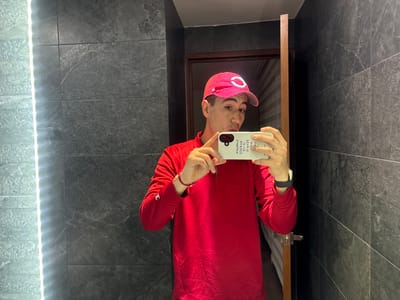A man is captured taking a mirror selfie indoors, likely in a bathroom, in San Bernardino Tlaxcalancingo, Mexico. He wears a red long-sleeved quarter-zip shirt and a red baseball cap with a prominent white "C" logo. He holds a white smartphone with his right hand, its screen obscured by his hand but showing text and camera details in reverse due to the mirror reflection. A black smartwatch is on his left wrist, and a red string bracelet adorns his right wrist.

The setting features dark grey, possibly marble or granite, tiled walls with subtle white veining. A bright vertical LED light strip runs along the left side of the mirror, illuminating the scene. In the reflection behind the man's right shoulder, a wooden door frame is visible, hinting at another room or hallway beyond. The man has a neutral to slightly pouting expression as he focuses on capturing the image.

Visible text on the back of the white phone case, appearing mirrored, includes phrases and words such as "SWIMMING," "SWIM," "WITH ME," "HI," "GE," "CE," and the number "3046." Additionally, standard camera lens labels like "ULTRA WIDE," "2X TELE," and "1X WIDE" are faintly discernible around the phone's camera module. The lighting is bright and artificial, suggesting an indoor setting.