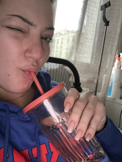 A young female is depicted indoors in Schiltigheim, France, from a frontal, chest-up perspective. She wears a blue hooded sweatshirt with red lettering and a thin gold-colored necklace. Her left eye is closed in a wink, while her right eye remains open and directed towards the viewer. She is actively drinking from a transparent, ribbed plastic tumbler with an orange-red lid and matching straw, holding it with her right hand. The tumbler contains liquid and displays exterior condensation. Her fingernails are manicured with a French tip style. The background includes a bright light source, consistent with a window covered by light-colored sheer curtains. A dark metal bed frame or chair back is partially visible to her left. To the right, a black extendable pole, resembling a selfie stick or stand, is present, alongside a partial view of a white storage unit holding various bottles.