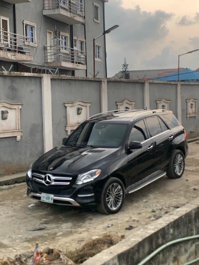 A black Mercedes-Benz GLE SUV, bearing Nigerian license plate "LKL-408JN" (Lekki, Lagos), is parked on a dirt and gravel surface next to a concrete drainage ditch in Lekki, Nigeria. The vehicle faces slightly left, its front bumper and grille visible, reflecting the overcast sky. A multi-story residential building, painted grey with multiple white-framed windows and balconies featuring metal railings, stands behind a tall, grey concrete perimeter wall topped with razor wire. The wall incorporates decorative rectangular recesses, each containing a solar-powered wall light. Two streetlights with LED fixtures are visible: one attached to the residential building's wall and another further in the background. A conical evergreen tree and a blue tarp-covered roof are visible over the perimeter wall. No individuals are present in the scene. Debris, including a plastic bottle, is scattered near the drainage ditch in the foreground. The sky is largely overcast with grey clouds.