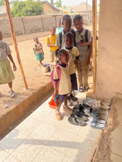 The image shows a group of school-aged children gathered outdoors, appearing to be in a line or congregation outside a building. The image is oriented vertically, with the ground surface appearing primarily on the right side and stretching towards the bottom, while the children and buildings occupy the left and central parts of the frame.
In the immediate foreground, on the lower right, there is a patterned tiled floor where several pairs of shoes, sandals, and slippers are neatly arranged against a light-colored wall. Many of these are black, while some are lighter in color, and a few are still wrapped in clear plastic. This arrangement suggests footwear might be removed before entering the adjacent building.
To the left of the tiled area, the ground transitions to a dirt or sandy surface. On this surface, a group of children is standing.
The closest child in this group, positioned centrally, is a young girl wearing a cream-colored top with pink sleeves and blue shorts, holding a small orange bucket or container. She is looking towards her left. Her feet are in light brown shoes.
Beside her, slightly to her left, is another young girl with braided hair, wearing a white collared shirt (possibly a school uniform with a visible logo on the left chest) and dark trousers. She is looking directly at the camera.
Behind these two, several other children are visible. One boy wears a similar white collared shirt and dark trousers, also looking towards the camera. Further back and slightly to the left, another boy in a grey collared shirt and light trousers is also looking at the camera. A young boy in a yellow short-sleeved shirt stands further back in the group. In the far background, a child in a light blue shirt is partially visible.
At the very bottom left of the frame, a person (likely an adult) is partially visible, wearing a light green skirt and a white top.
The background consists of concrete block walls of buildings and some distant sparse vegetation, suggesting a semi-urban or rural setting. A rough wooden beam or pole is lying horizontally on the dirt ground in the lower-left portion of the image.
The scene is brightly lit by strong sunlight, casting distinct shadows on both the tiled floor and the dirt ground, indicating a clear, sunny day, likely morning or late afternoon given the angle of the shadows. The children generally have neutral or slightly curious expressions. No text is visible in the image.