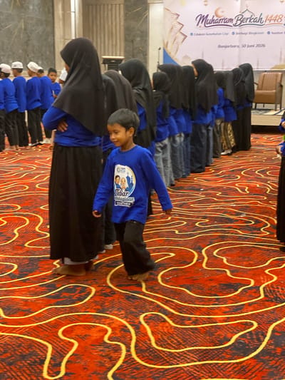 The image shows a group of children, mostly boys, gathered indoors on a red carpet with a yellow wavy pattern. The children are wearing blue long-sleeved shirts and dark pants or skirts. Some of the girls are wearing black headscarves. In the background, a banner reads "Muharram Berkah 1448 H" and mentions "Banjarbaru, 30 Juni 2026," indicating the location and date of an event, likely a religious celebration or gathering. Some adults, also wearing headscarves, are present in the group. The lighting suggests an indoor event in a hall or auditorium. One boy in the foreground is walking towards the viewer and appears to be smiling. The overall scene depicts a community event for children in Banjarbaru, Indonesia.