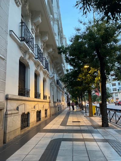 The image shows a street scene in Madrid, Spain, near the intersection of Calle de Serrano and Calle de Goya. A pedestrian sidewalk runs alongside a white building with decorative balconies and large windows. Trees line the sidewalk, casting shadows on the patterned pavement. A street lamp illuminates the area. Several people are walking on the sidewalk, and cars are visible on the street. The sky is a clear blue, suggesting it is daytime.
